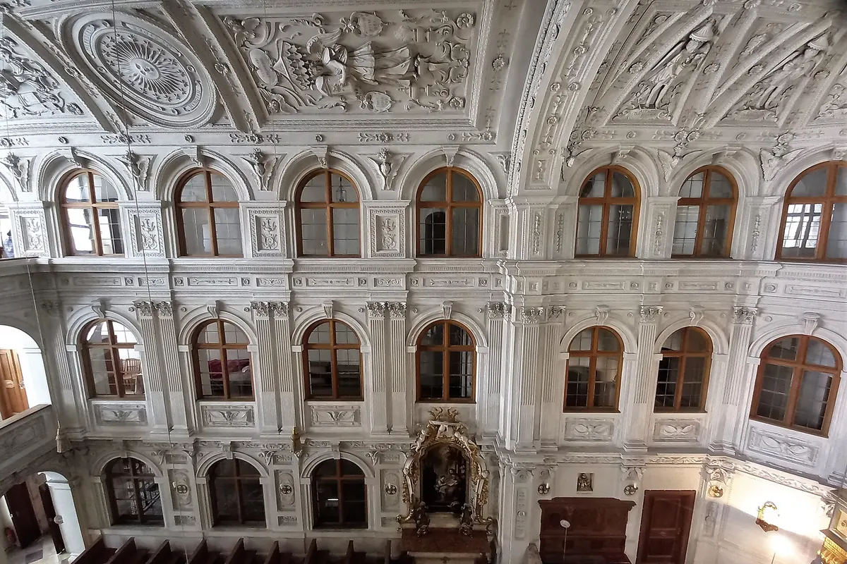 The upper wall of the chapel with two storeys of arched windows, stucco panels and wooden pews below.