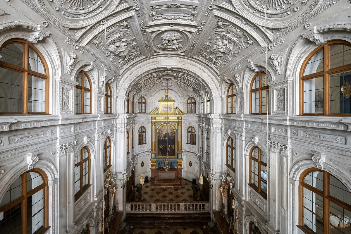 The court chapel seen from the gallery, with a tall white stucco nave, arched windows with brown frames and a gilded altar at the far end.